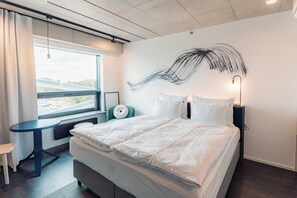 A modern hotel room with a large bed, a small table, a chair, and a view of the outside through a window.