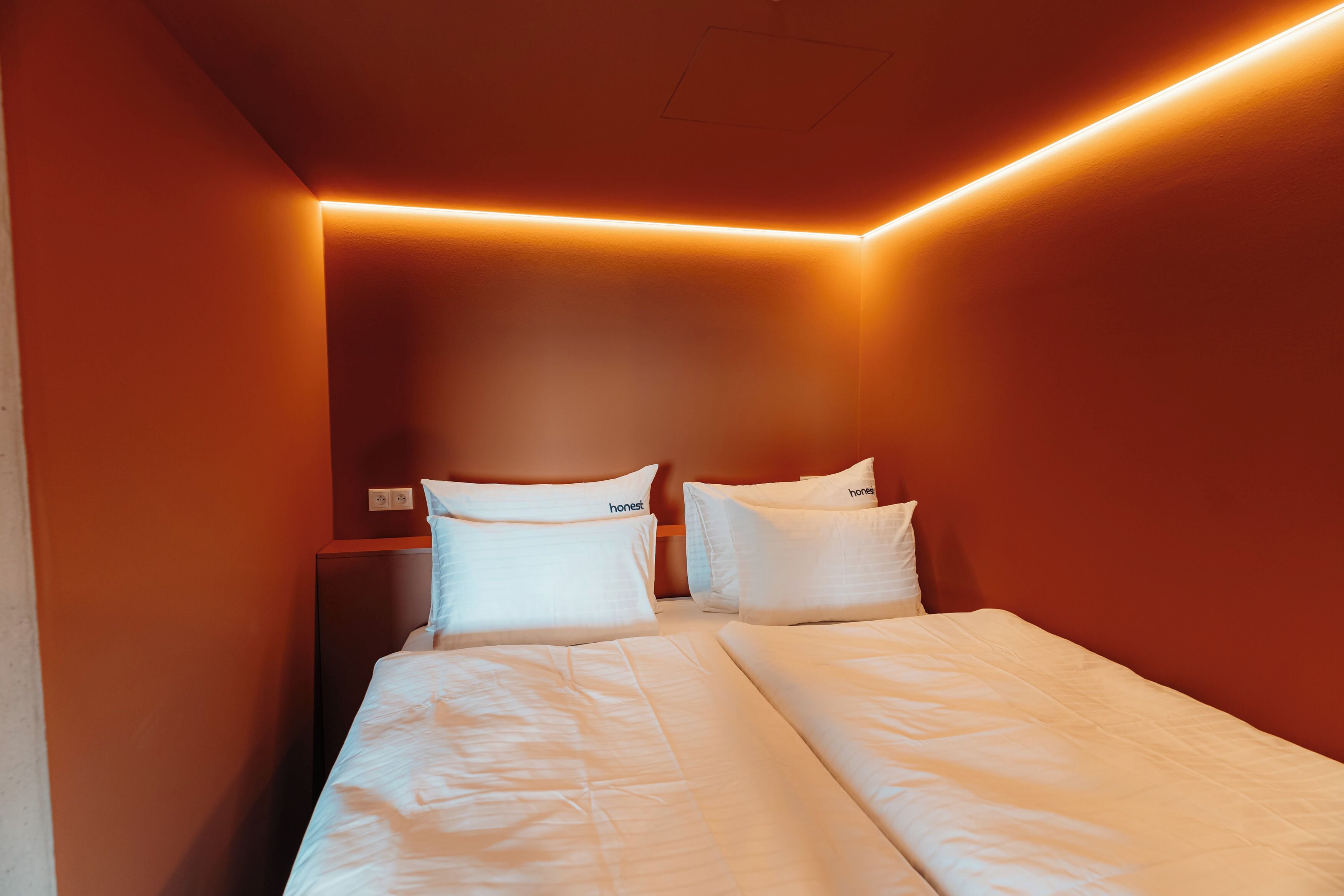 Quarto de hotel com duas camas, roupa de cama branca e uma parede cor de laranja quente.