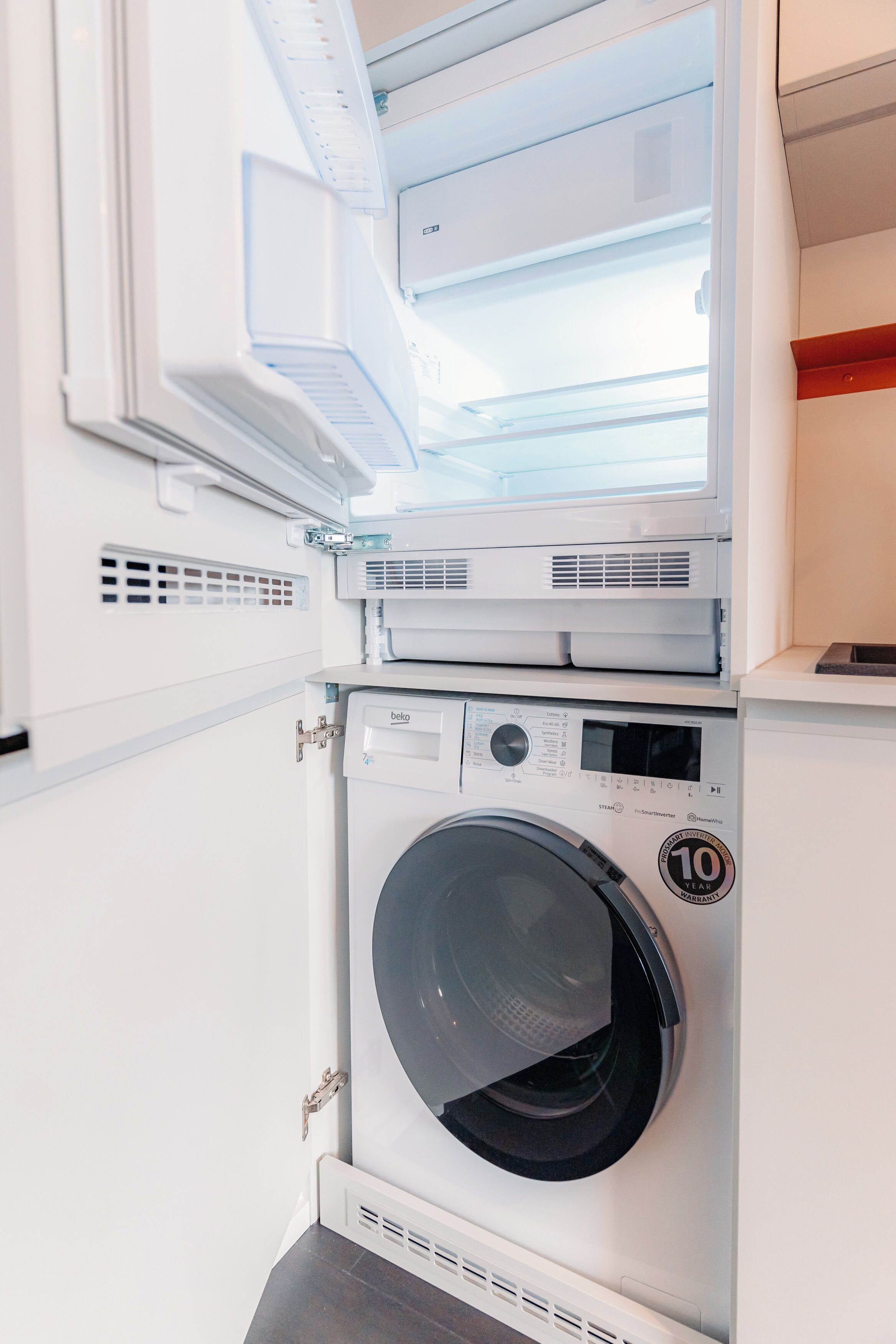 Une petite buanderie moderne, dotée d’un lave-linge et d’un sèche-linge à chargement frontal, d’une petite fenêtre au-dessus du sèche-linge et d’un sol carrelé blanc.