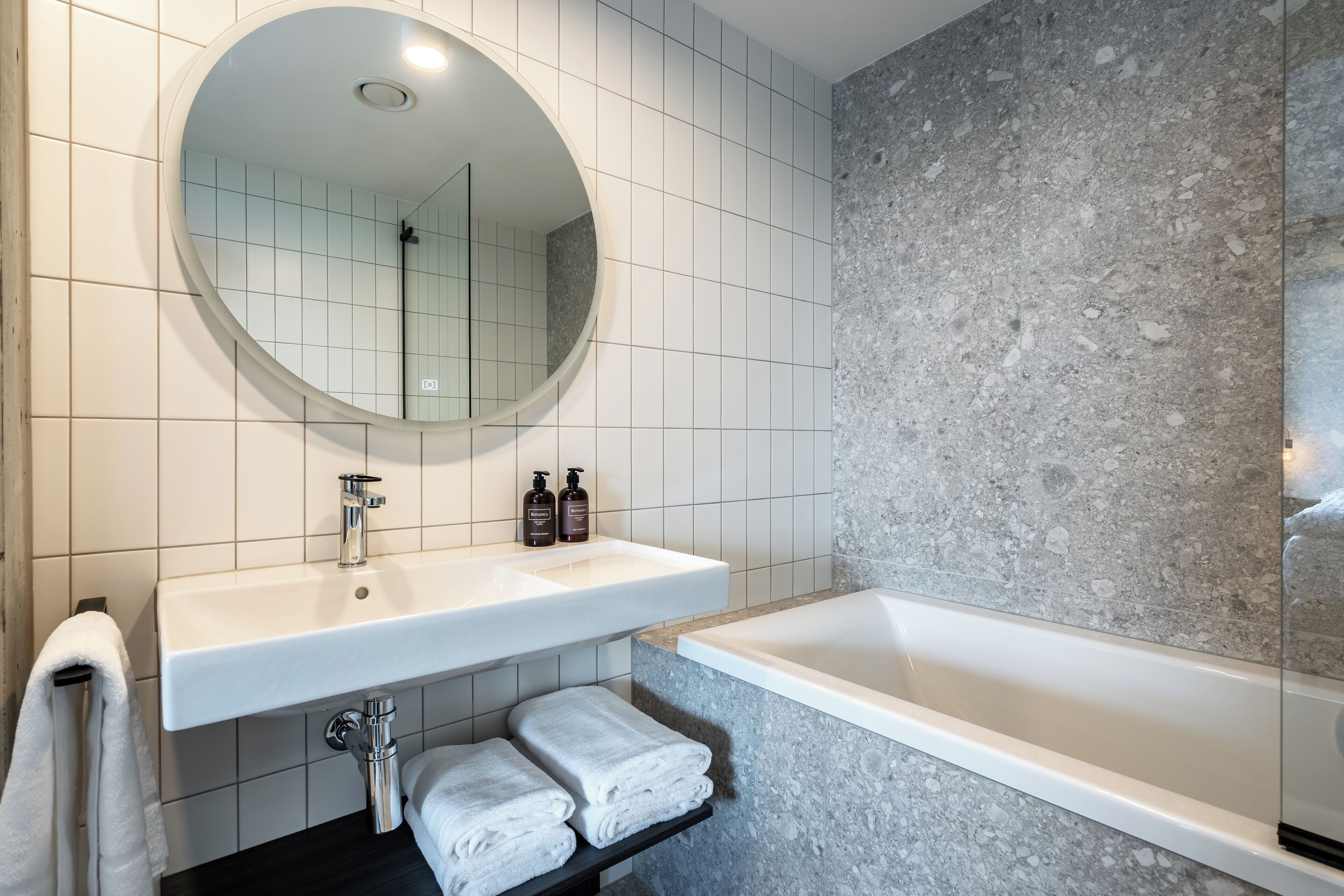 Ein modernes Badezimmer mit rundem Spiegel, zwei Waschbecken, einer Badewanne und ordentlich gefalteten Handtüchern.