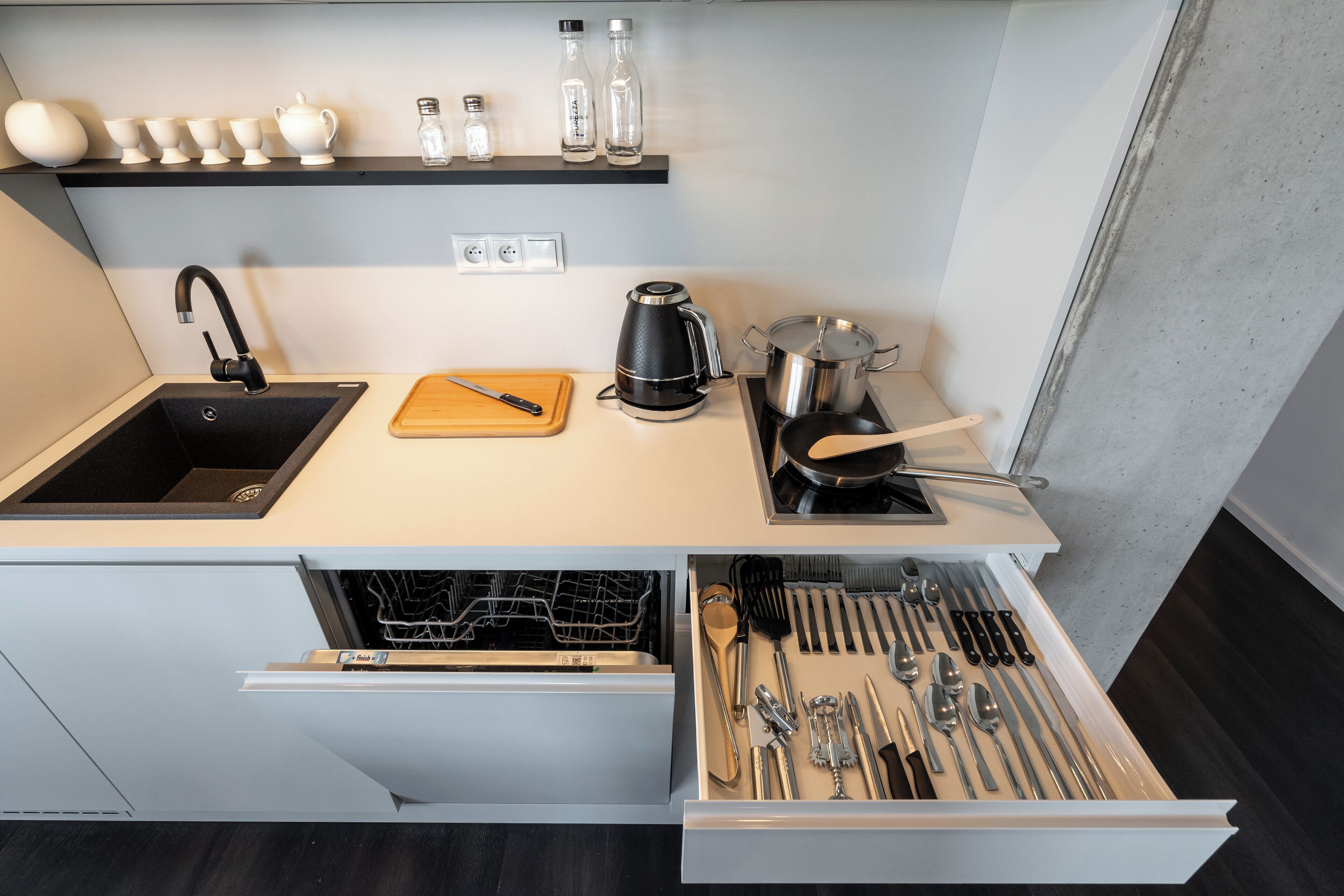 Una cocina moderna con un lavamanos negro, una cafetera, una tabla de cortar y un cajón con utensilios.