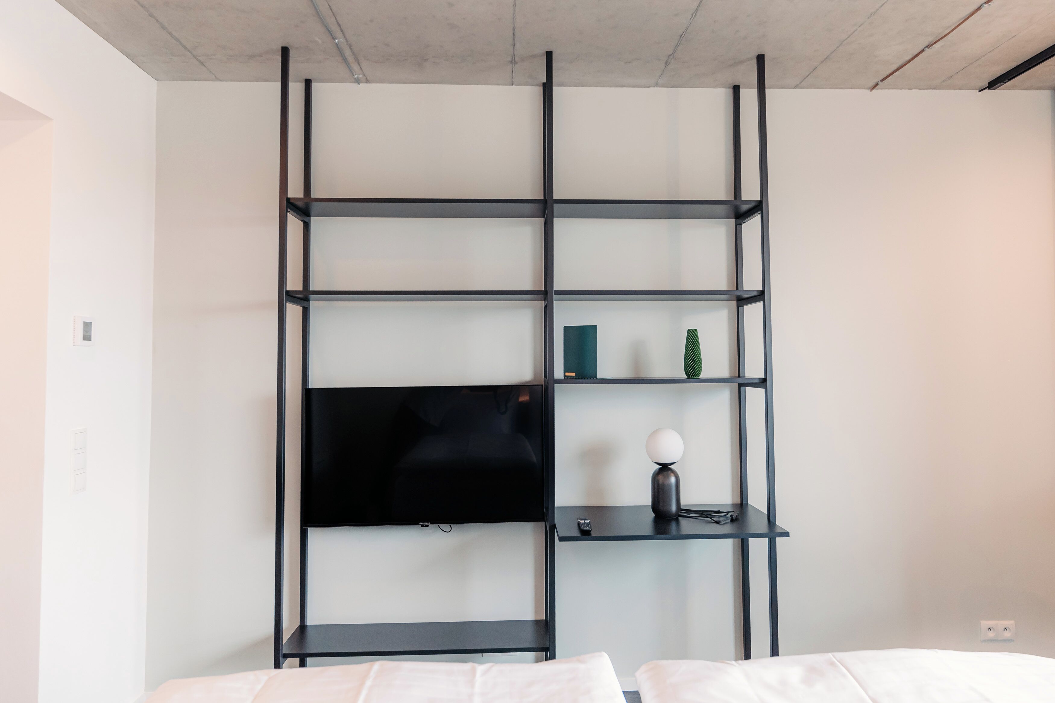 A modern room with a black TV on a wall-mounted shelf, a small table with a lamp, and a bed with white bedding.