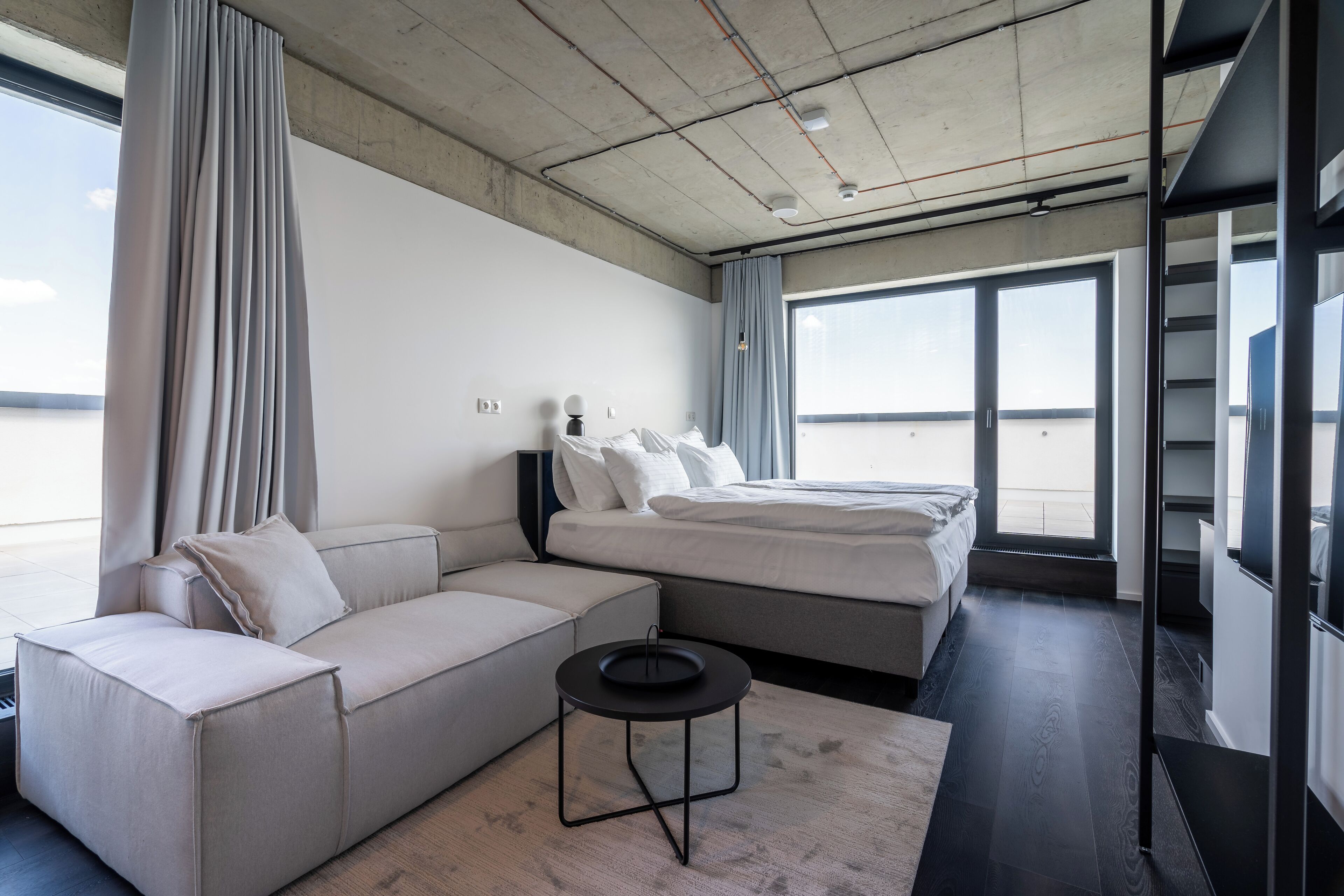 A modern hotel room with a large bed, a sofa, a small table, and a view of the sea through large windows.