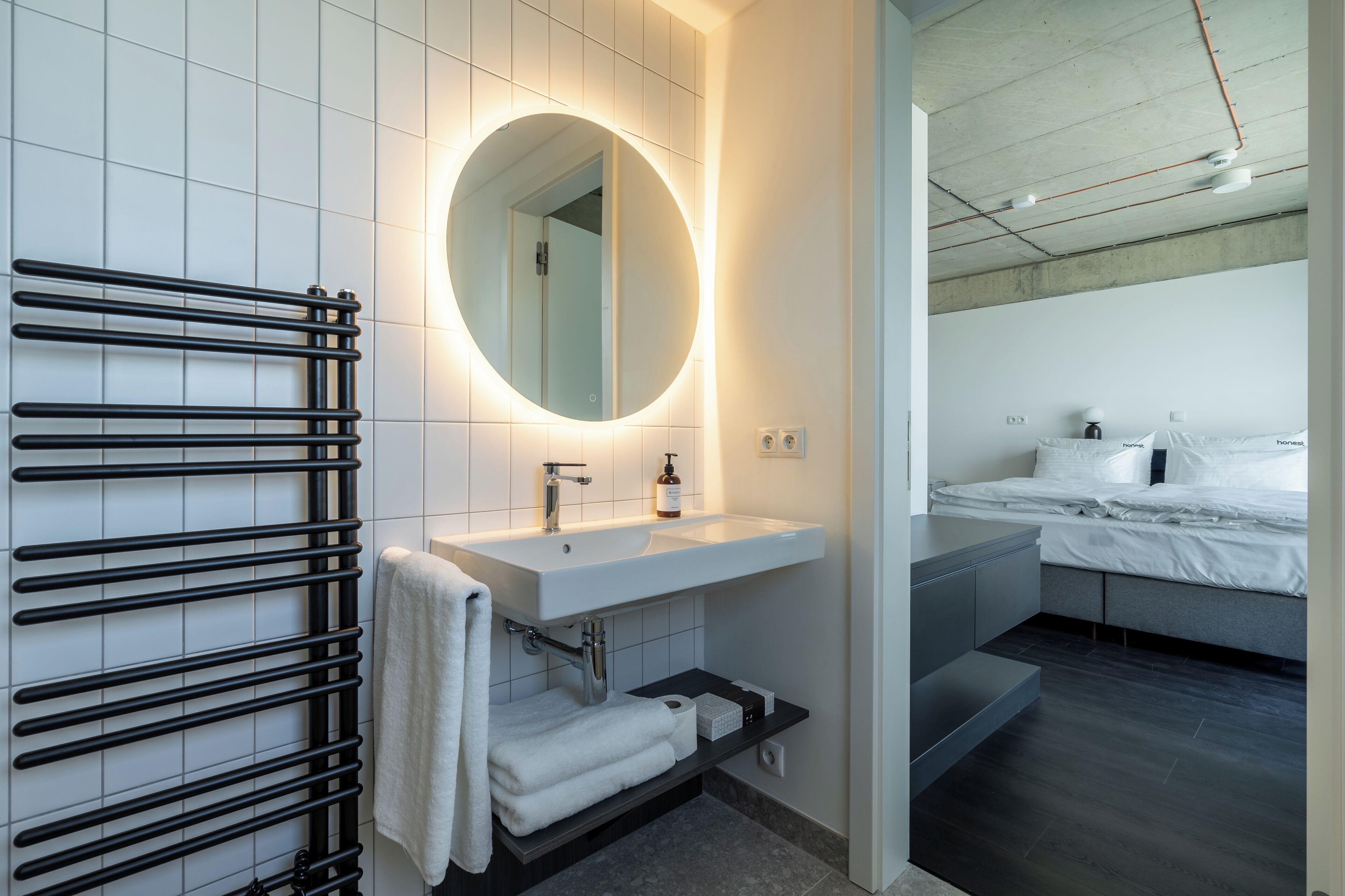 A modern bathroom with a black towel rack, a round mirror, a white sink, and a bed with white bedding.