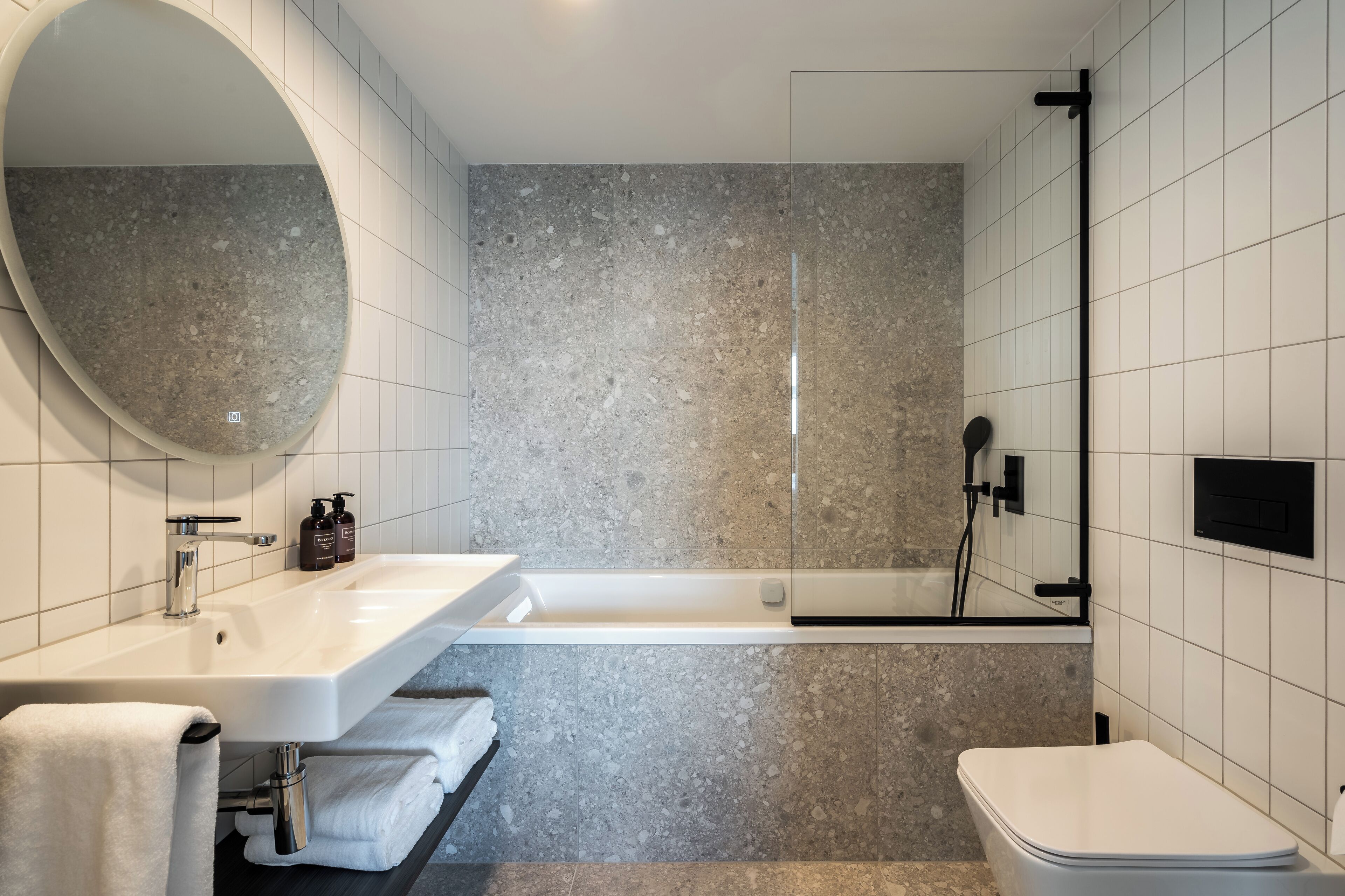 A modern bathroom with a large oval mirror, a white sink, a bathtub, and a toilet.