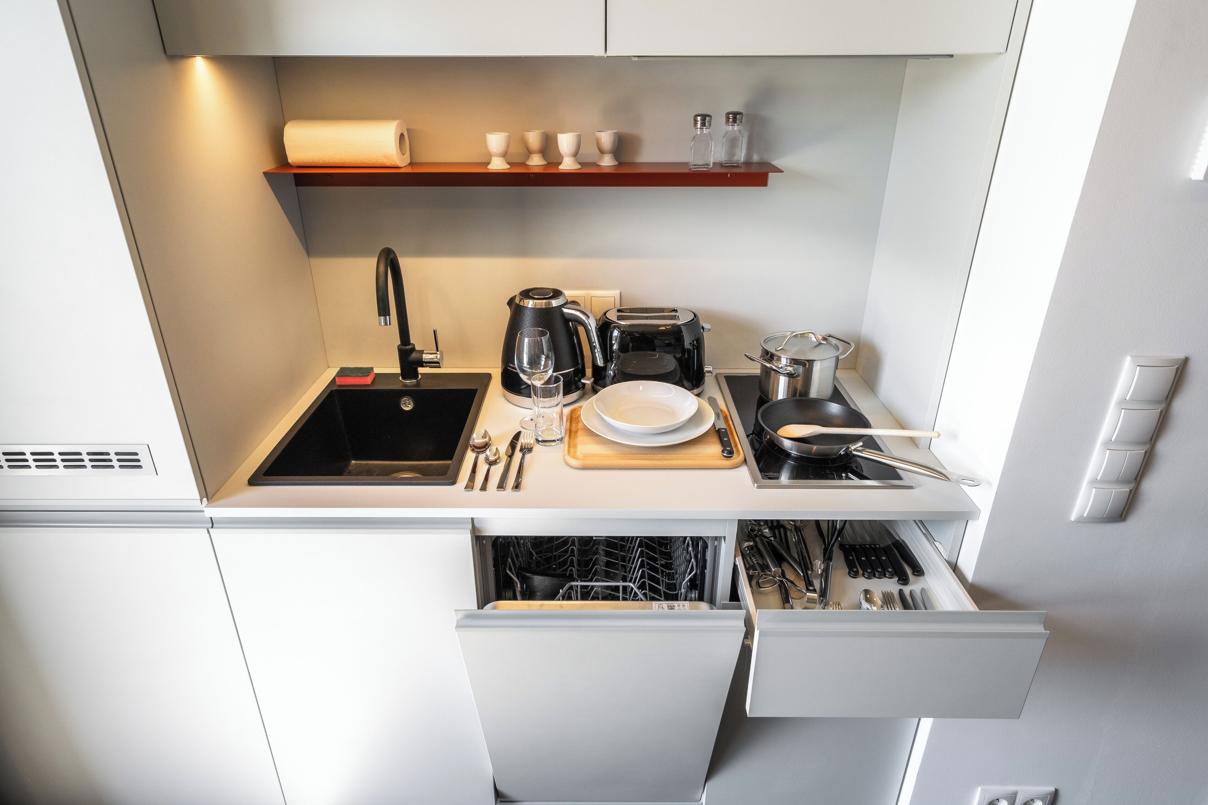 A modern kitchen with a sink, stove, and dishwasher. There are utensils, a coffee maker, and a cutting board on the counter.