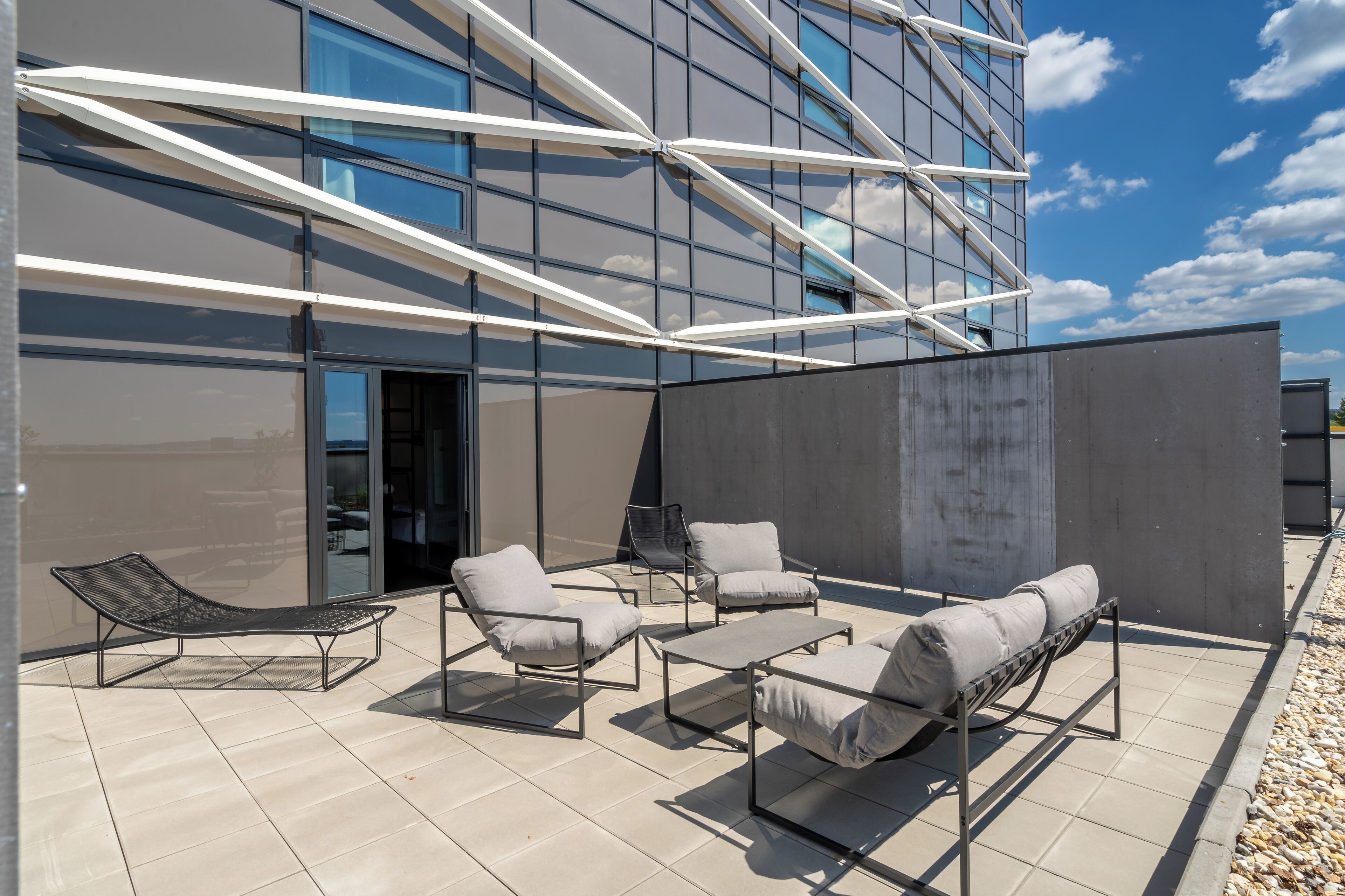 An outdoor seating area with modern furniture, a glass building facade, and a clear sky.
