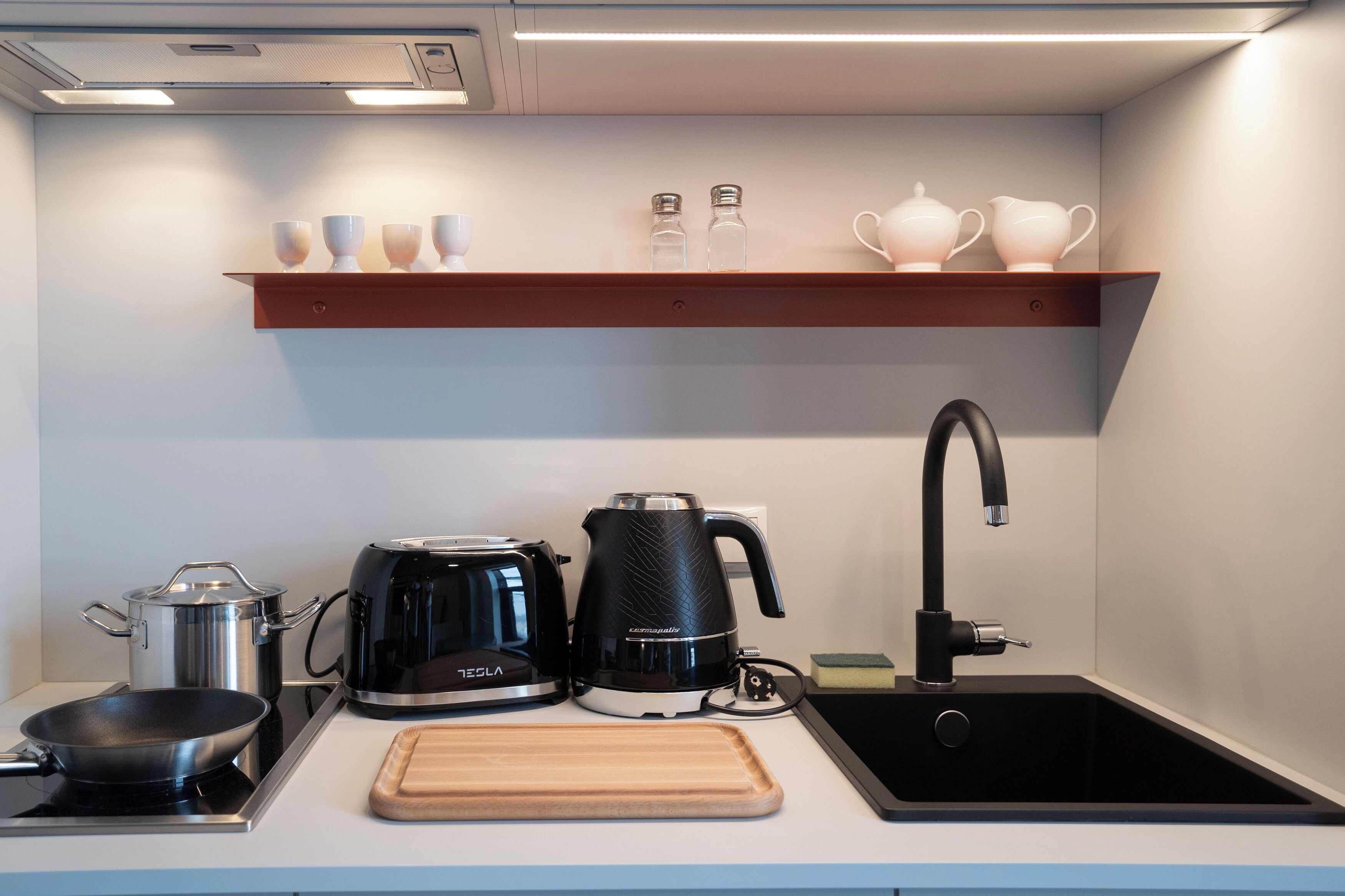 Een kleine keuken met een kookplaat, een koffiezetapparaat, een waterkoker, een spoelbak en een houten snijplank.