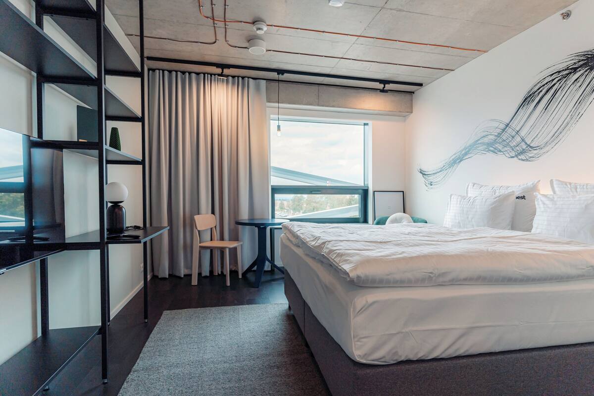 A modern hotel room with a large bed, a desk, and a chair. There is a window with curtains, a wall art piece, and a view of the outside.