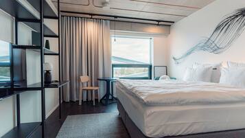 A modern hotel room with a large bed, a desk, and a chair. There is a window with curtains, a wall art piece, and a view of the outside.