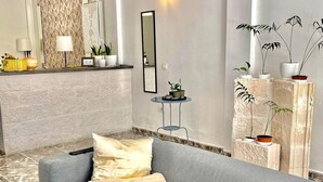 A modern living room with a grey sofa, two cushions, a small round table with a lamp, and a wall-mounted mirror. There are several potted plants on a white stone shelf and a white wall with a textured pattern. - Spacious loft in the heart of a quiet and lively beach town.  (Arona)