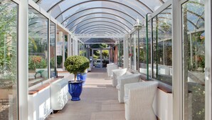 A covered walkway with glass walls, white wicker furniture, and potted plants.