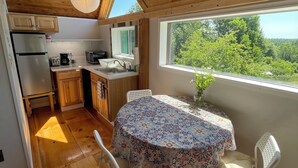 A wooden interior with a round table, chairs, a kitchen area, and a window overlooking greenery. - Cozy Cottage (Surry)