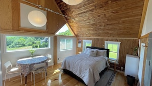 A cozy bedroom with a wooden ceiling, a large bed, a dining table with chairs, and a view of greenery outside. - Cozy Cottage (Surry)