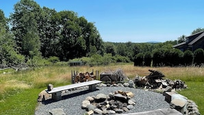 A circular fire pit surrounded by rocks and logs, with benches and a small shed in a grassy field. - Cozy Cottage (Surry)