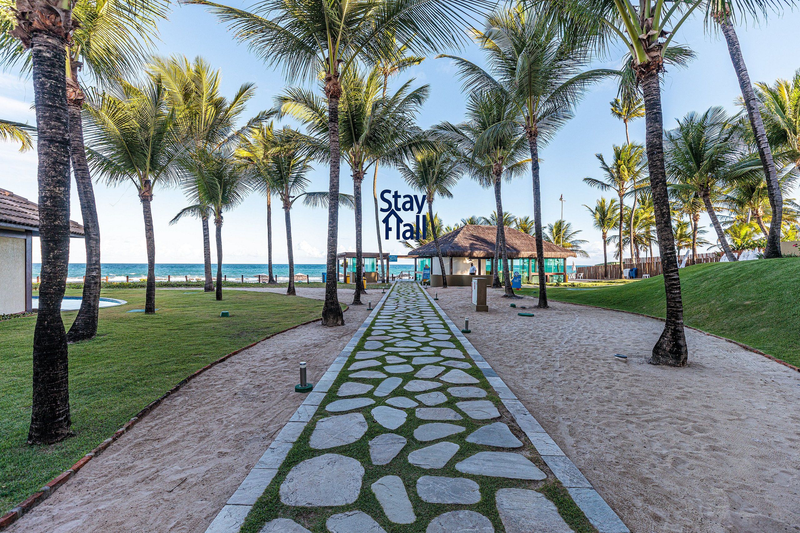 A pathway lined with palm trees leading to a building with a thatched roof, surrounded by a sandy area and a grassy lawn.