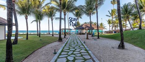 A pathway lined with palm trees leading to a building with a thatched roof, surrounded by a sandy area and a grassy lawn.