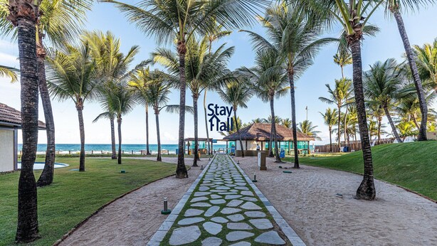 A pathway lined with palm trees leading to a building with a thatched roof, surrounded by a sandy area and a grassy lawn. - Bangalô 4qts BeiraMar em Muro Alto-SH034 (Ipojuca)