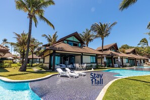 A resort with a swimming pool, lounge chairs, palm trees, and a 'Stay Here' sign. - Bangalô 4qts BeiraMar em Muro Alto-SH034 (Ipojuca)