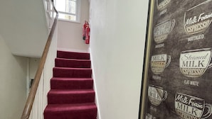A staircase with red carpeted steps and a handrail, leading to a landing with a fire extinguisher and a window. - Oban Victoria Hotel (London)
