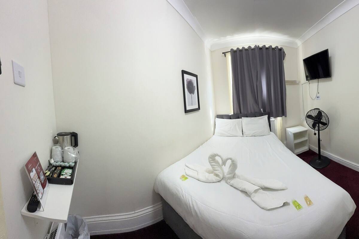 A hotel room with a neatly made bed, a bedside table with a remote control, a fan, and a TV on the wall.