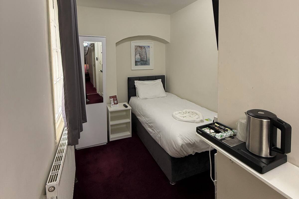 A compact hotel room with a single bed, a bedside table with a kettle, a small shelf, and a radiator.