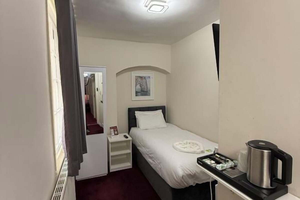 Una habitación de hotel pequeña y ordenada, con una cama individual, una mesita de noche con una tetera y un bandeja de artículos de aseo, y un cuadro en la pared.