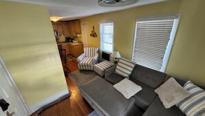 Un salón con un sofá seccional gris, sillones a rayas y una mesita. Se ve una cocina al fondo, con armarios de madera y una ventana.