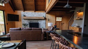 Ein gemütliches Wohnzimmer mit Kamin aus Stein, Holzwänden und einem großen Fernseher. Im Hintergrund befindet sich eine Bar mit Hockern und eine Küche.