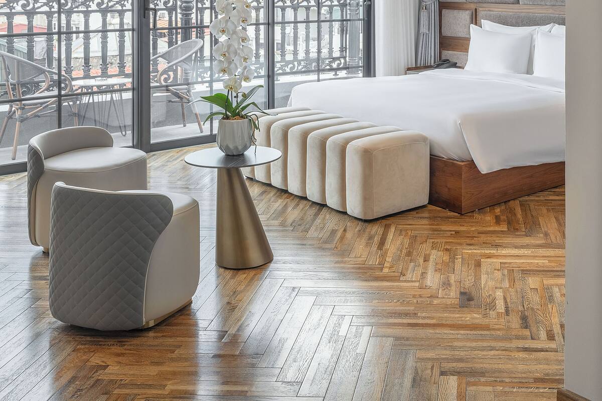 Una habitación de hotel moderna con una cama grande, un área de descanso con otomanas, una mesita y vistas de la ciudad a través de amplios ventanales.