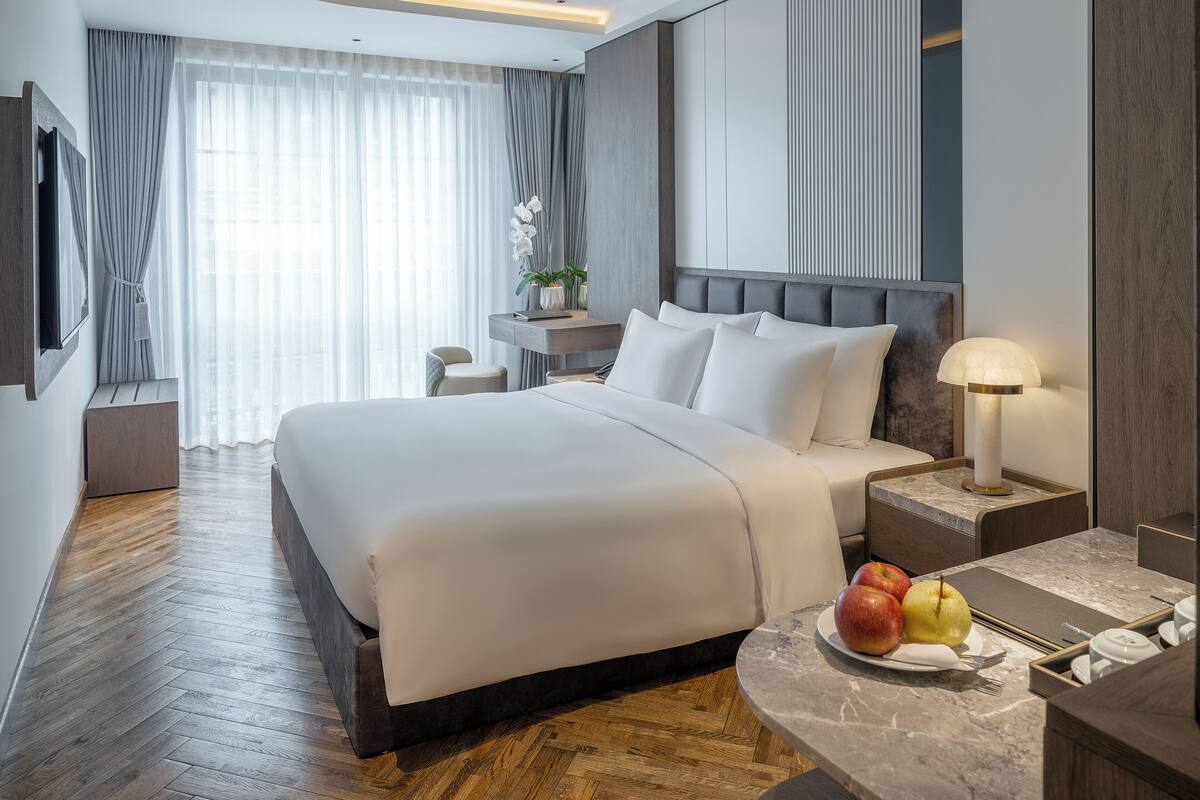 Una habitación de hotel moderna con una cama grande, una mesita de noche, una lámpara y un frutero en la mesita.