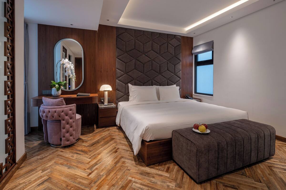 Un dormitorio moderno con suelo de parqué, una cama grande con ropa blanca, una mesita de noche con lámpara, un tocador con espejo y una silla tapizada en rosa.