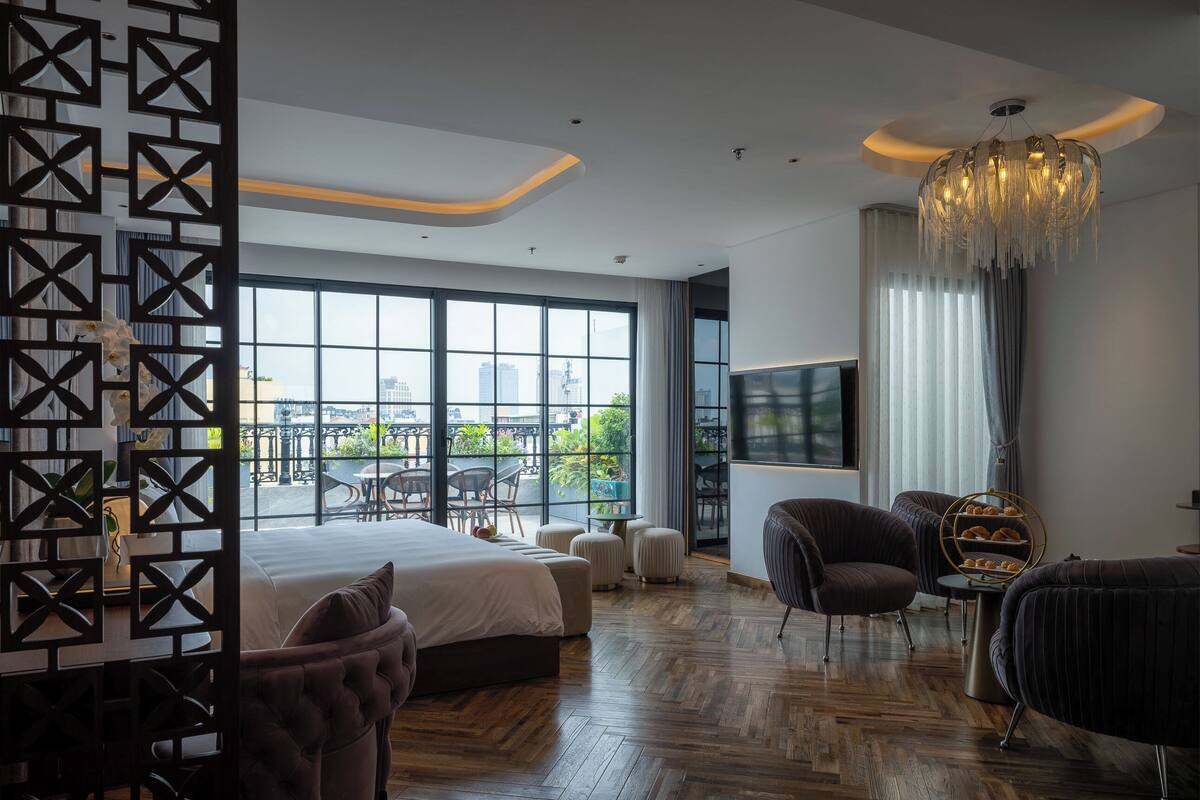 Una habitación de hotel moderna con una cama grande, televisión, zona de estar con sillones y vistas a la ciudad a través de amplias ventanas.