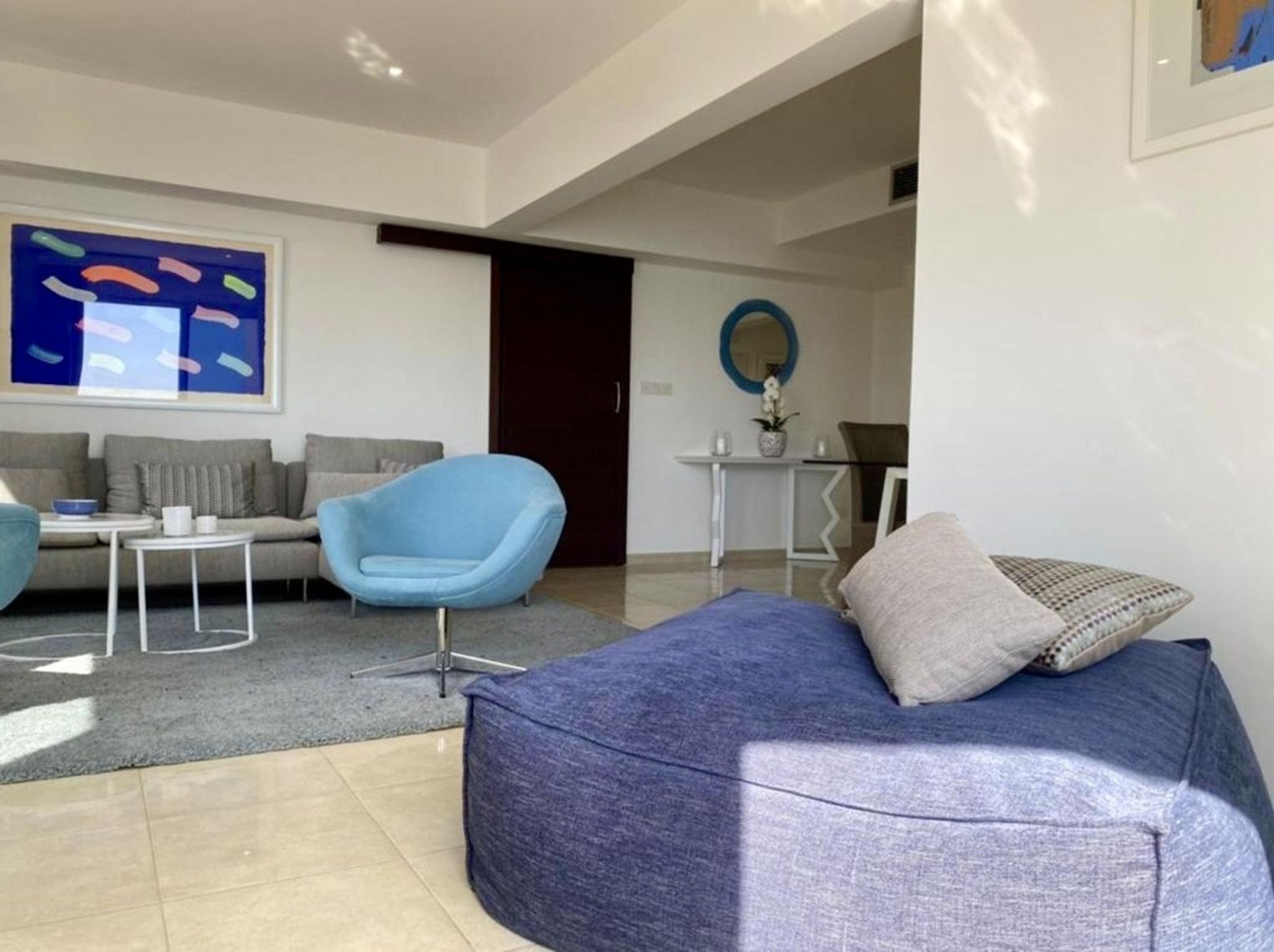 Una sala de estar moderna con un sofá azul, un sillón azul claro y un sofá gris. Hay una pequeña mesa blanca con un tazón y una taza. Un cuadro abstracto grande está en la pared, y un espejo redondo sobre una mesa blanca.