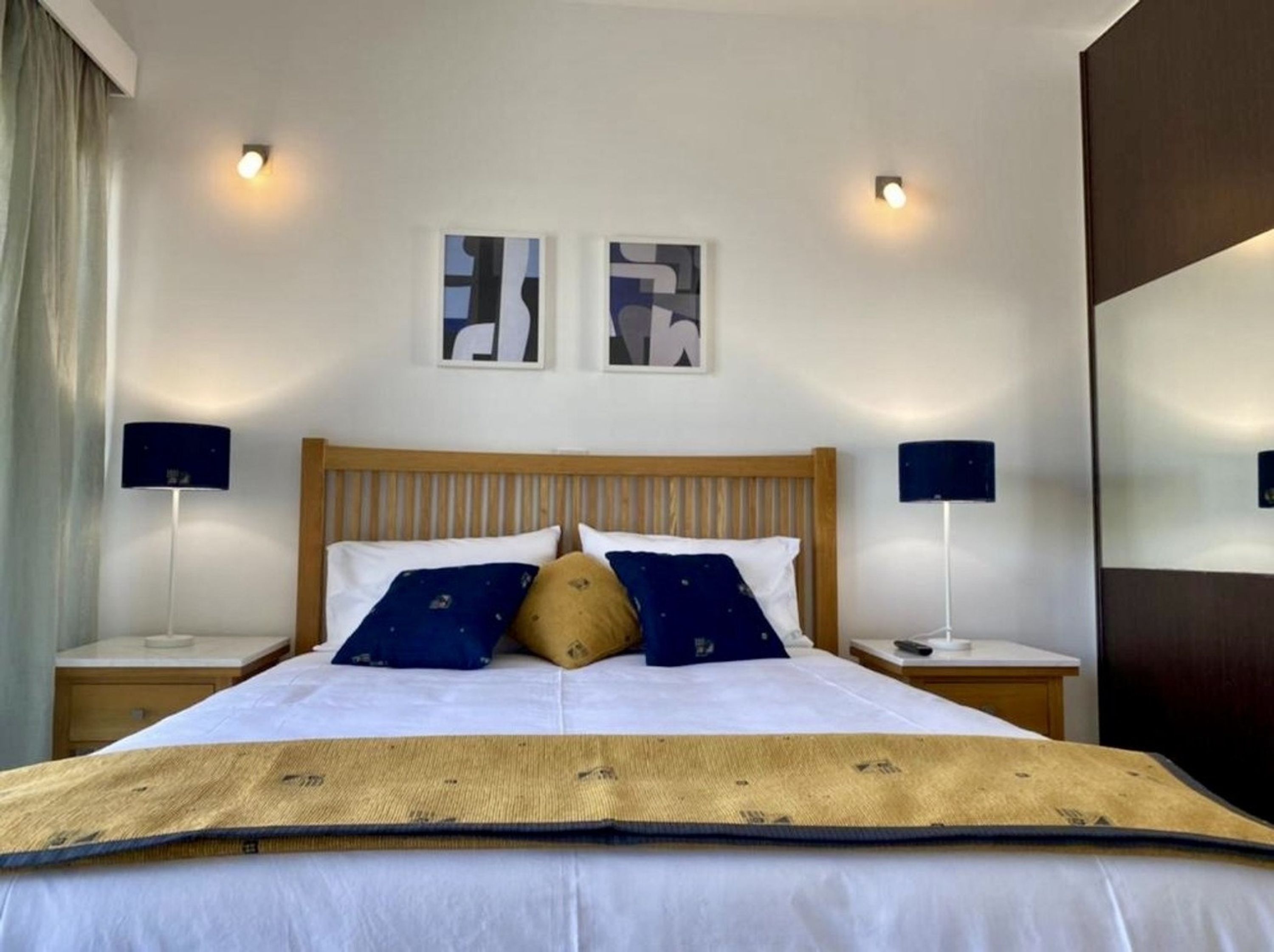 A hotel room with a wooden bed, two bedside tables, a large mirror, and abstract wall art.