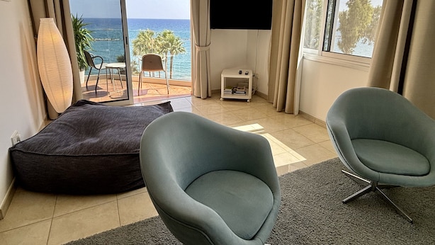 Una habitación moderna con un amplio ventanal corredero que da a un balcón con vistas al mar. Hay dos sillas azul claro, una mesita auxiliar blanca con un control remoto y una lámpara de pie grande.