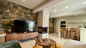 Een moderne woonkamer met een stenen muur, een flatscreen televisie, een open haard en een eethoek, met een keuken op de achtergrond.