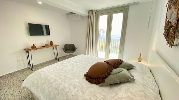 Een slaapkamer met een groot bed, een bureau, een stoel, een televisie en een balkon met gordijnen.