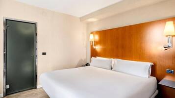 Una habitación de hotel con una cama grande, cabecera de madera, dos lámparas fijadas a la pared y una puerta.