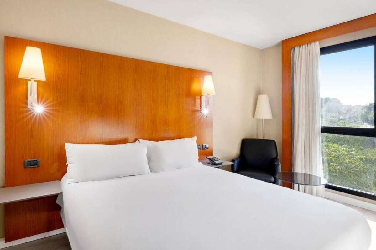 A hotel room with a large bed, two wall-mounted lamps, a chair, a small table, and a large window with a view.