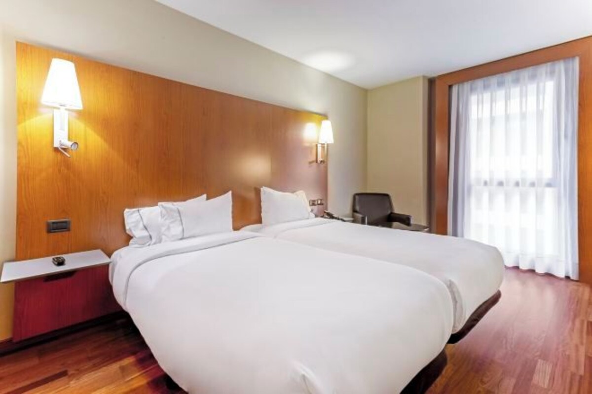 A hotel room with two beds, wooden headboard, bedside lamps, a desk, and a window with curtains.