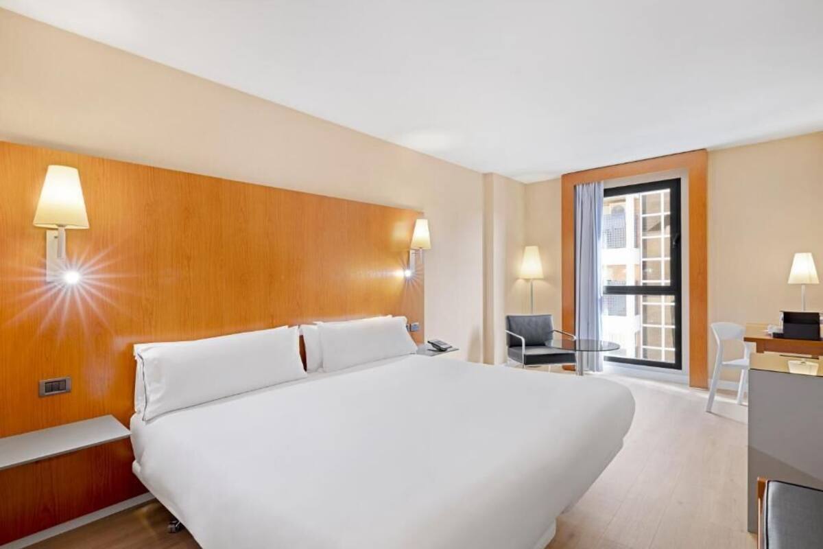 A hotel room with a large bed, bedside lamps, a desk with a chair, and a view of the outside through a glass door.