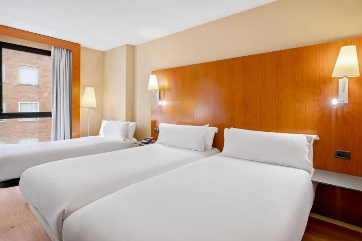 A hotel room with two beds, wooden headboard, bedside lamps, and a window with curtains.