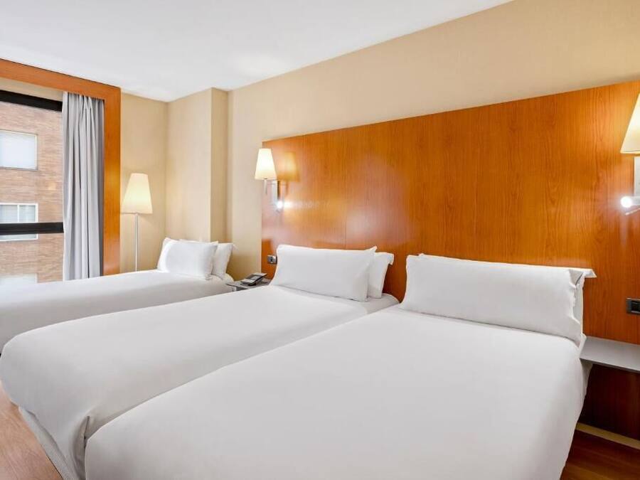 Una habitación de hotel con dos camas, cabecero de madera, lámparas de noche y una ventana con cortinas.