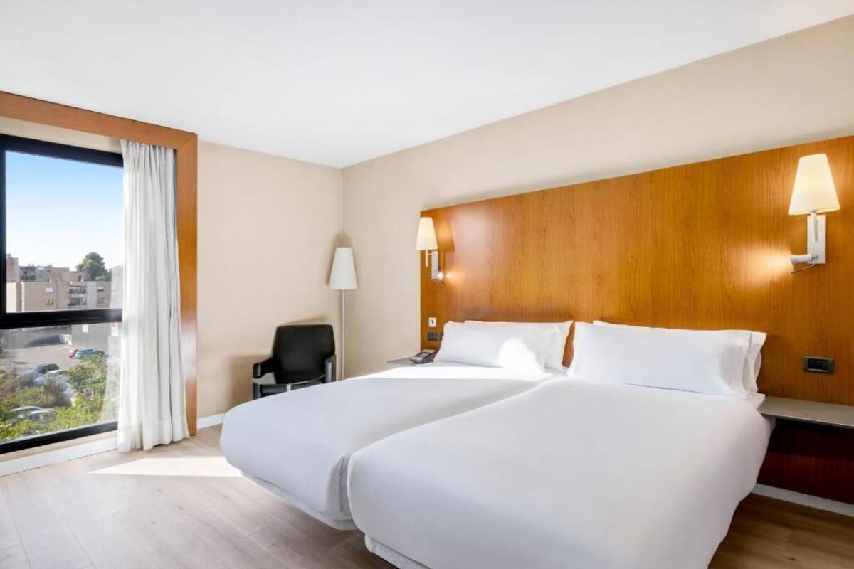 A hotel room with two beds, a wooden headboard, a desk, a chair, and a window with a view of buildings.
