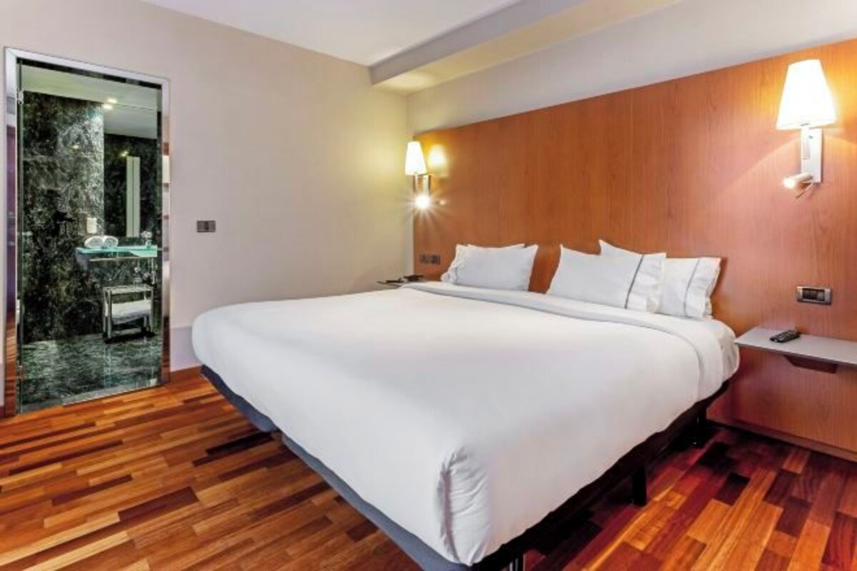 A hotel room with a large bed, wooden flooring, a glass shower enclosure, and wall-mounted lamps.