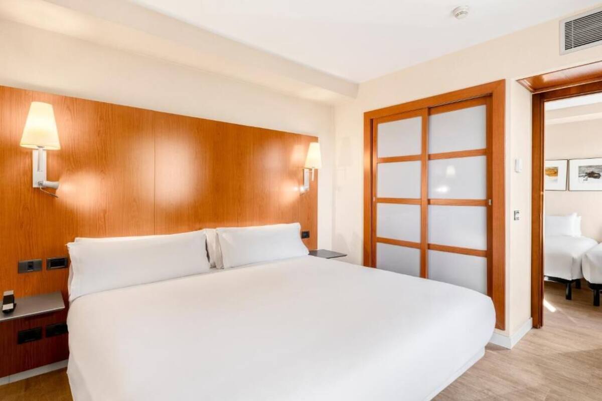 A hotel room with a large bed, wooden headboard, two bedside lamps, a nightstand, and a door with frosted glass panels.
