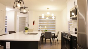 A modern kitchen with a central island, stainless steel appliances, and a dining area with a chandelier. - Brand New| 3 Bedroom, 1 king 2 Queen| Near Airport (Calgary)