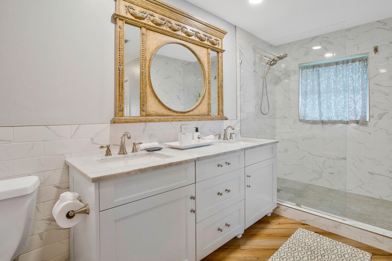 A bathroom with a marble shower, a large mirror with a gold frame, a white sink, and a toilet.