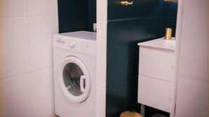 A compact laundry room with a front-loading washing machine, a small trash bin, and a white cabinet. - Modern Apartment with Sunny Terrace and Sea View (La Possession)